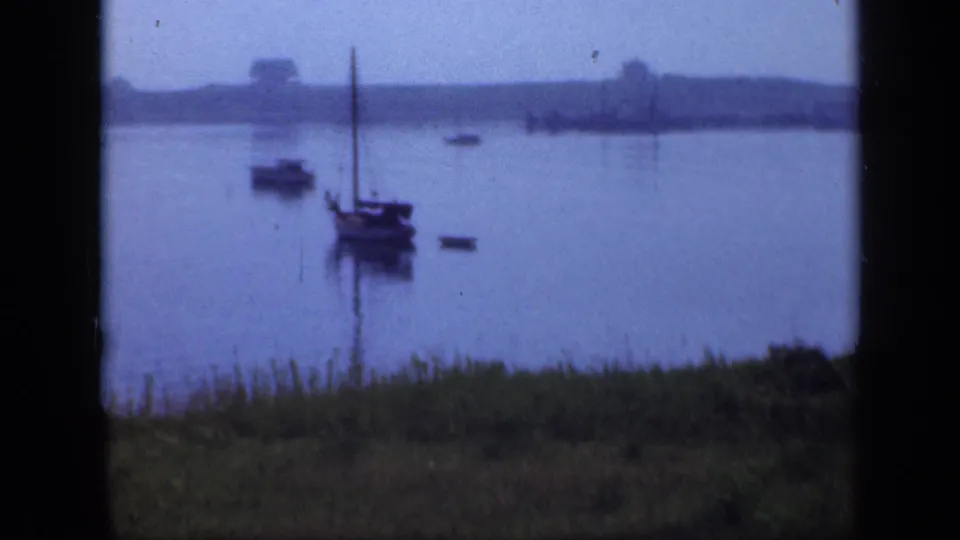 Soft 8mm frame of a moored sailboat on a still Long Island lake, with grass in foreground and low shoreline beyond.