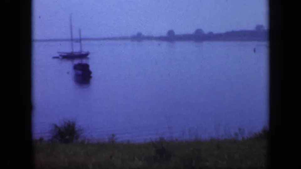 1948 Long Island waterfront frame with sailboats and a small dark boat resting on calm water near the shore.