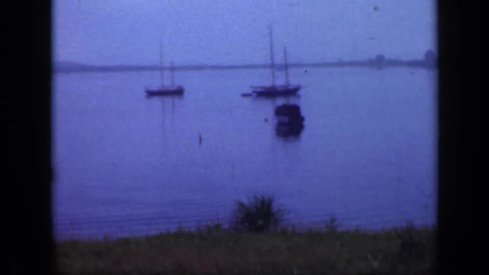 Vintage 8mm Long Island harbor view with anchored sailboats, a small boat, still water, and a low distant shore.