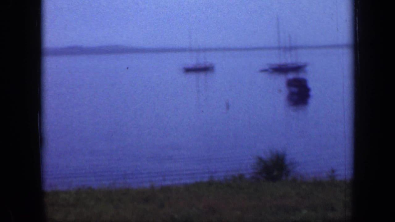 Anchored boats on calm water with a grassy foreground and dark film-frame edges.