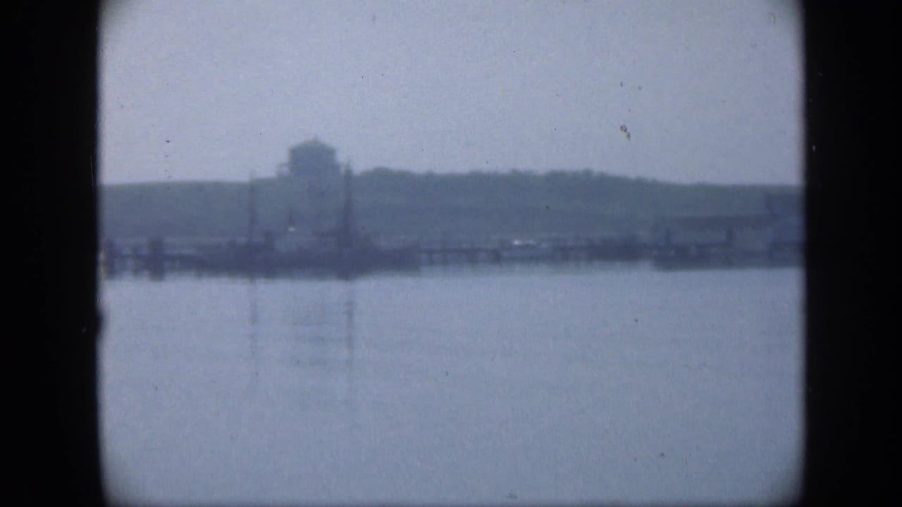 Hazy water view with distant shoreline, grassy rise, poles, and a small structure.