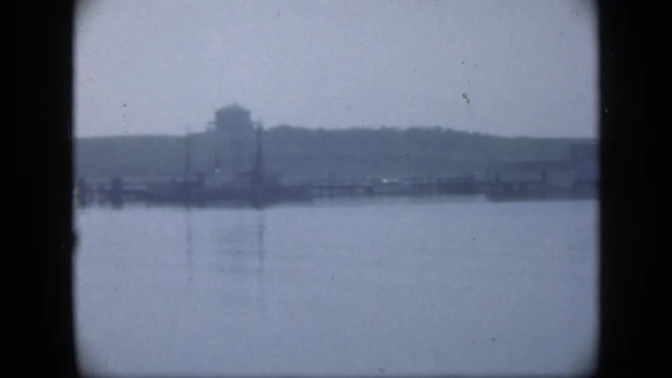 1948 Long Island 8mm frame looking across a broad body of water toward a hazy grassy shoreline with poles and a distant structure.