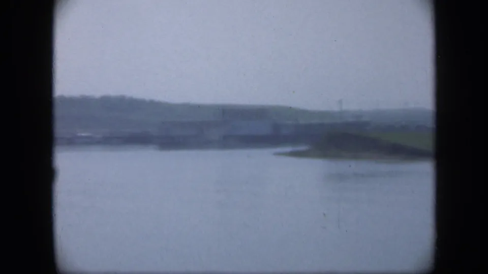 1948 Long Island home-movie frame showing a wide water surface and low grassy shoreline, with faint distant structures softened by haze.