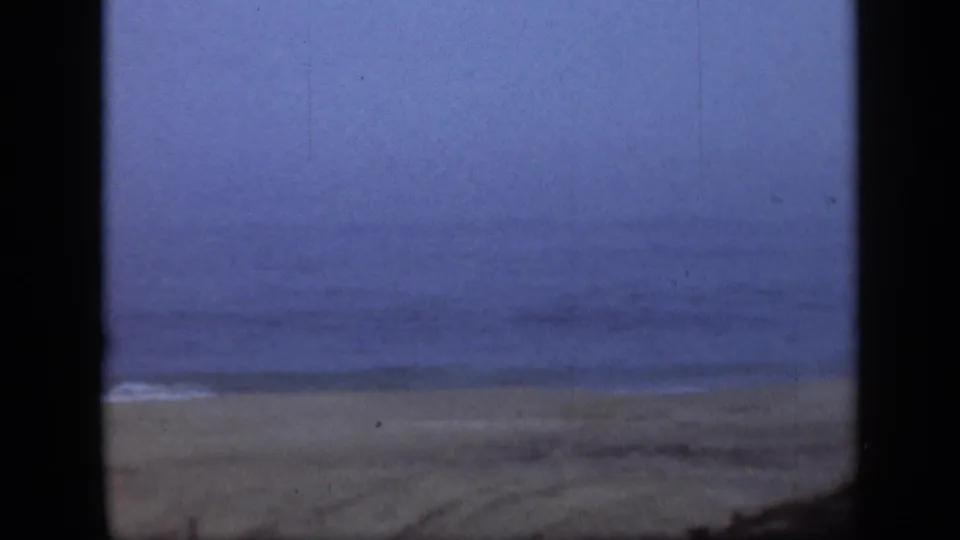 Wide 1948 Long Island shoreline view with sand, soft surf, blue sky, and heavy dark borders from the filming position.