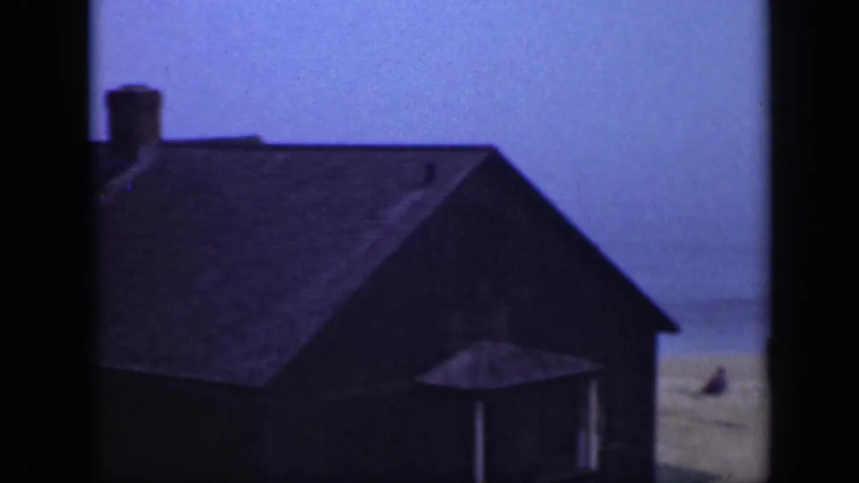 Dark sloped-roof building near the Long Island shoreline, with sand, surf, blue sky, and dark film-frame borders visible.