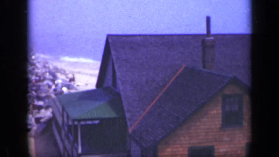 1948 Long Island coastal frame with a large shingled house, small green shed, rocky shoreline, and surf in the distance.