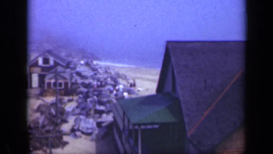 1948 Long Island home-movie frame showing beach houses clustered beside rocks, sand, surf, and a small green shed.