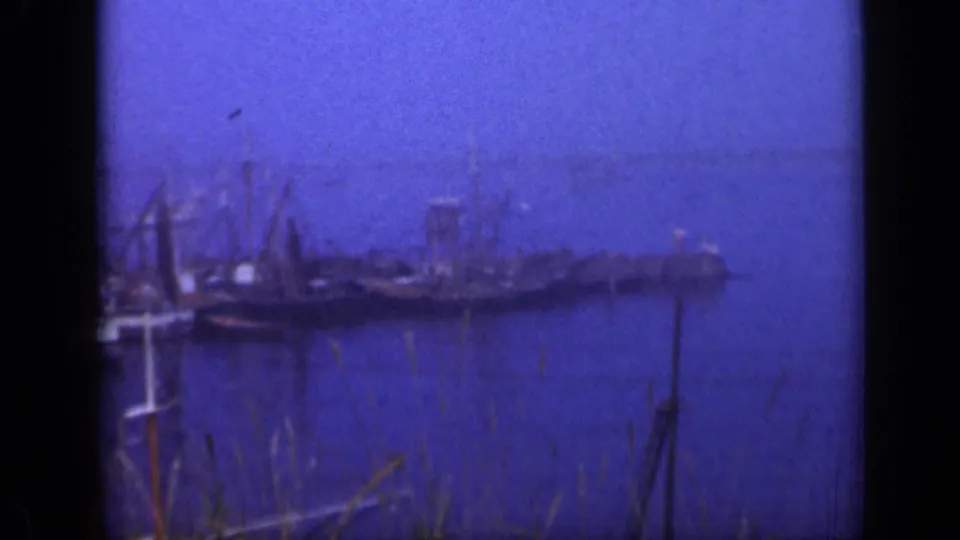 1948 Long Island waterfront frame showing boats at a pier, masts, shoreline grasses, calm water, and a hazy background shore.