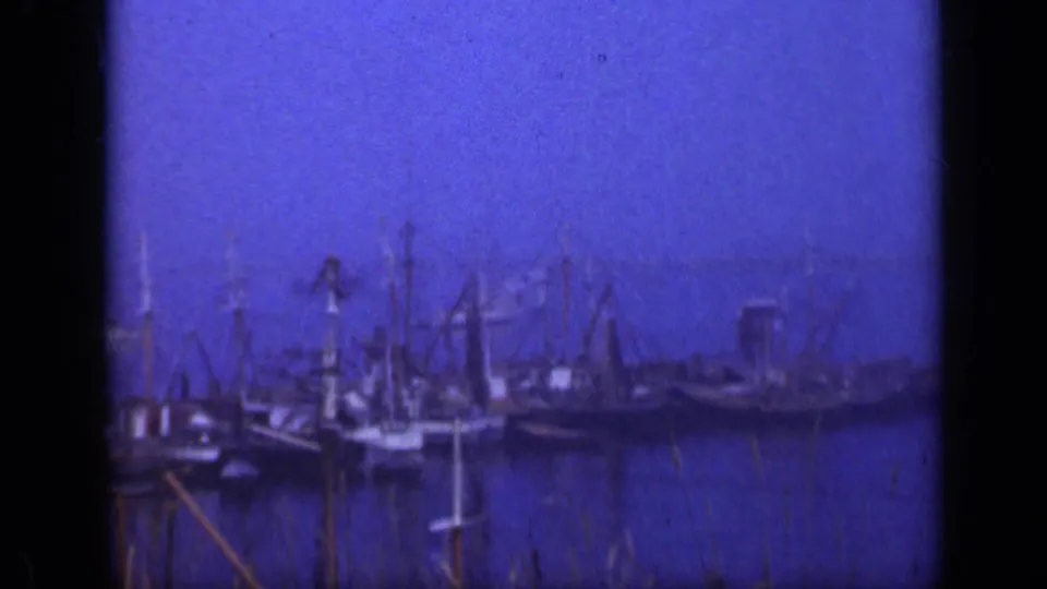1948 Long Island marina view with old sailboats, tall masts, rigging, dark water, and foreground grass in blue-tinted 8mm footage.