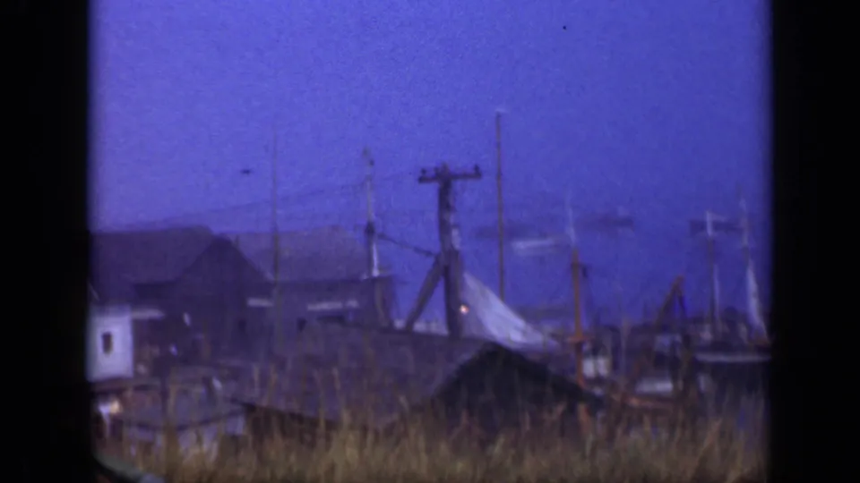 Long Island harbor-side buildings and utility poles appear beyond foreground grass and dock structures in this 1948 8mm frame.
