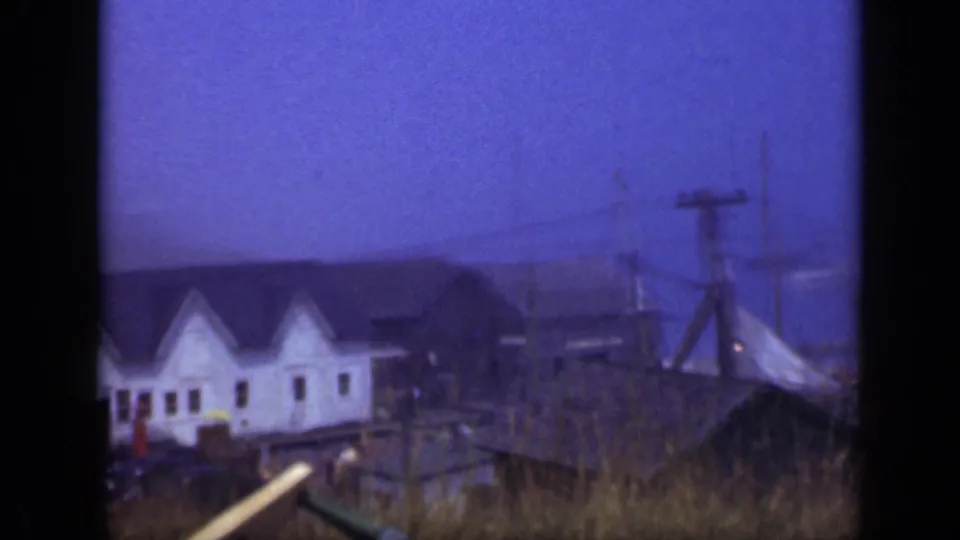 A row of white waterfront buildings sits near docks and utility poles in a soft 1948 Long Island home-movie view.