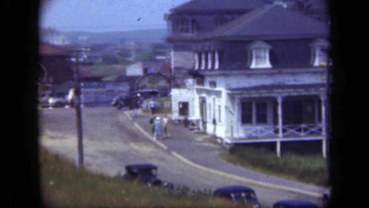 Street corner with a large white house, parked cars, utility poles, and distant pedestrians.