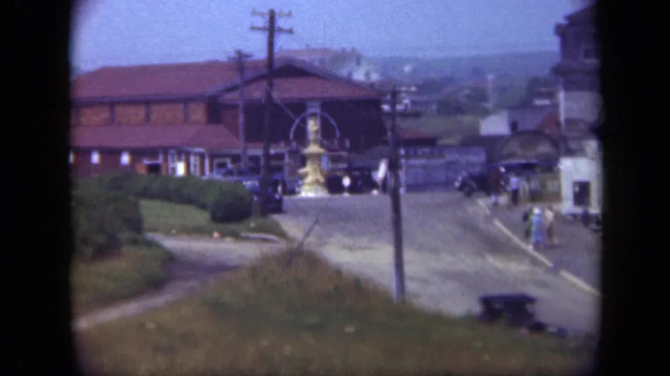 1948 Long Island town view showing a brick building, golden fountain, utility poles, parked automobiles, and distant pedestrians.