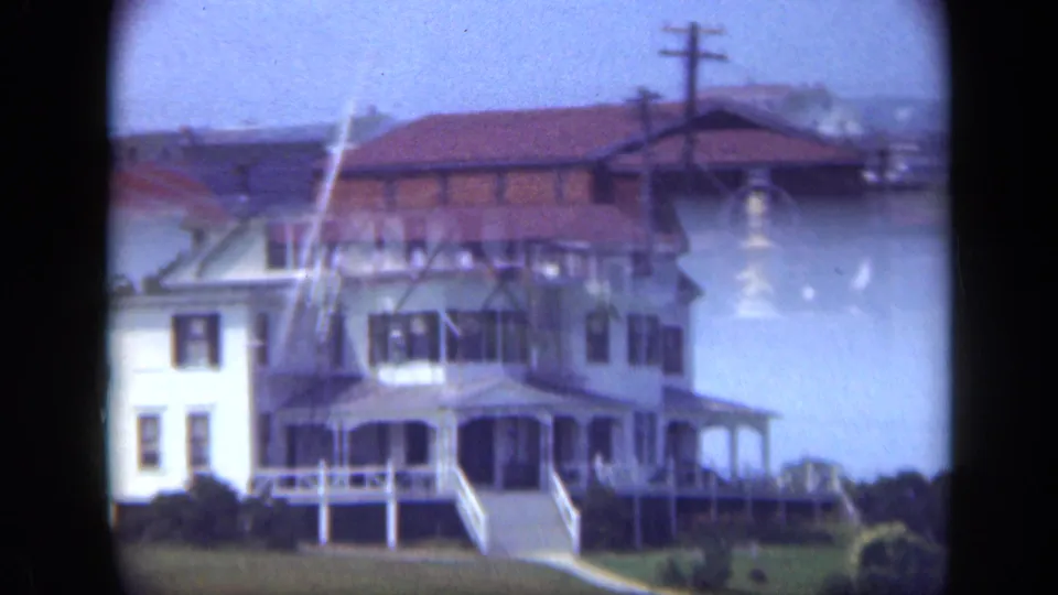 1948 Long Island exterior of a white Victorian-style house with porch, front steps, lawn, and a brick building in the background.