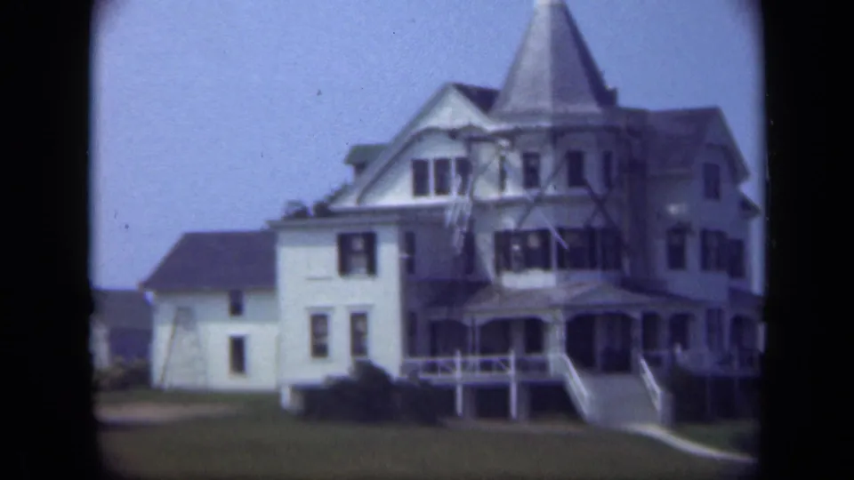 1948 Long Island home-movie frame showing a large multi-story house with steep rooflines, porches, front steps, and open lawn.
