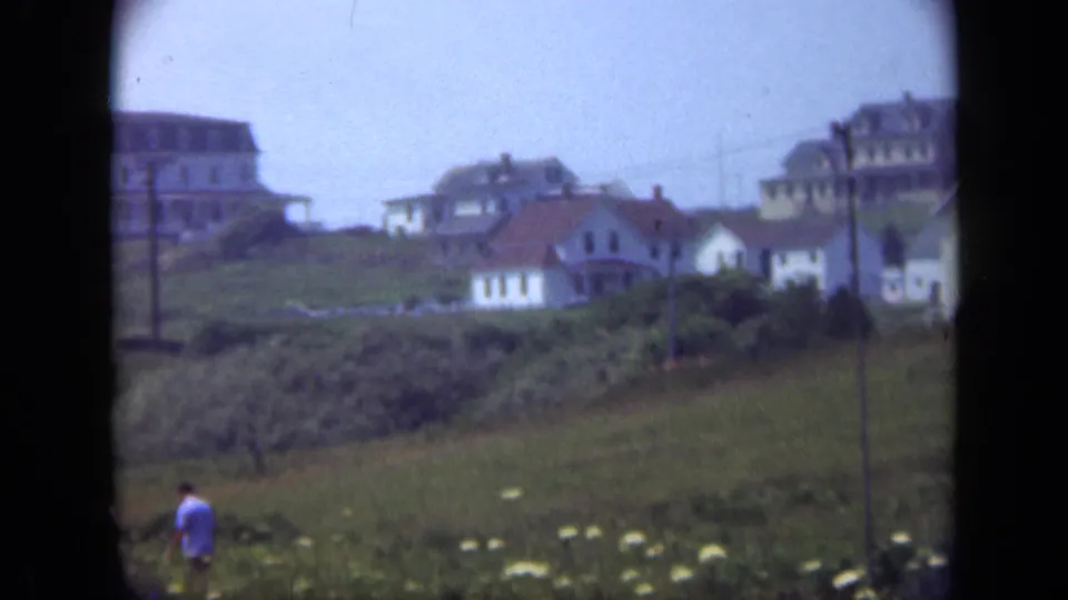 1948 Long Island home-movie view across a grassy hillside with several houses, utility poles, shrubs, and a distant walker.