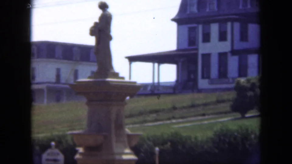 1948 Long Island frame showing a stone statue on a pedestal, lawn, shrubbery, and part of a large porch-fronted building.