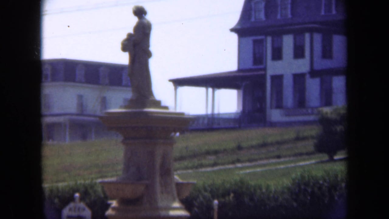 1948 Long Island 8mm frame showing a statue on a pedestal in front of a grassy field and large building.