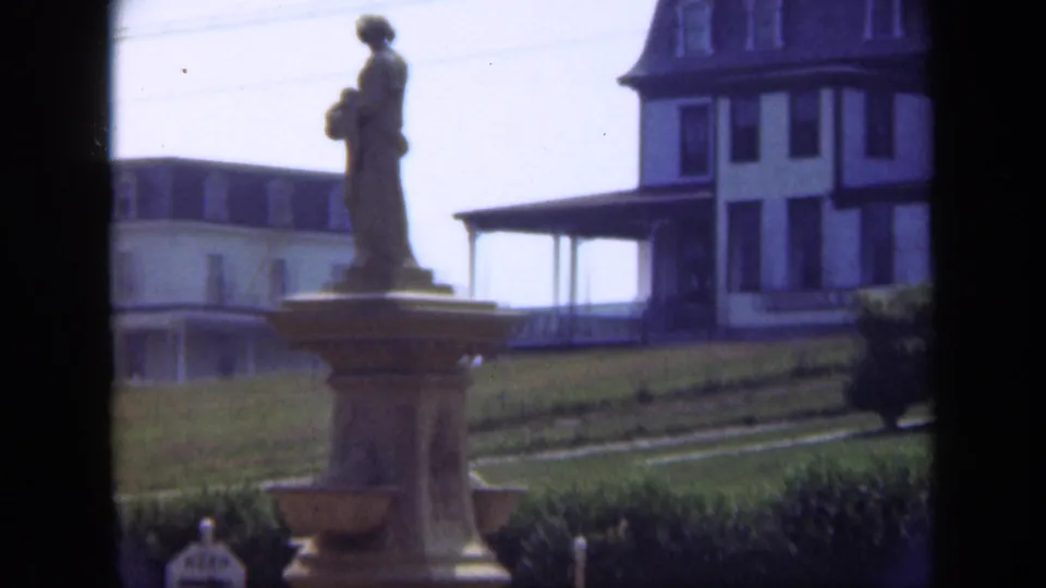 1948 Long Island 8mm frame showing a statue on a pedestal in front of a grassy field and large building.