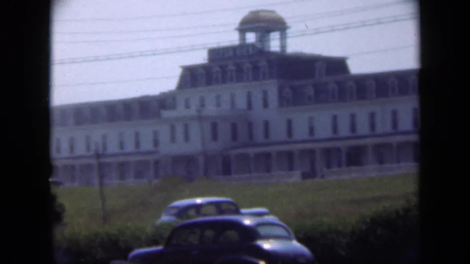 1948 Long Island 8mm view of a large building with a domed cupola, broad lawn, hedges, and parked cars.