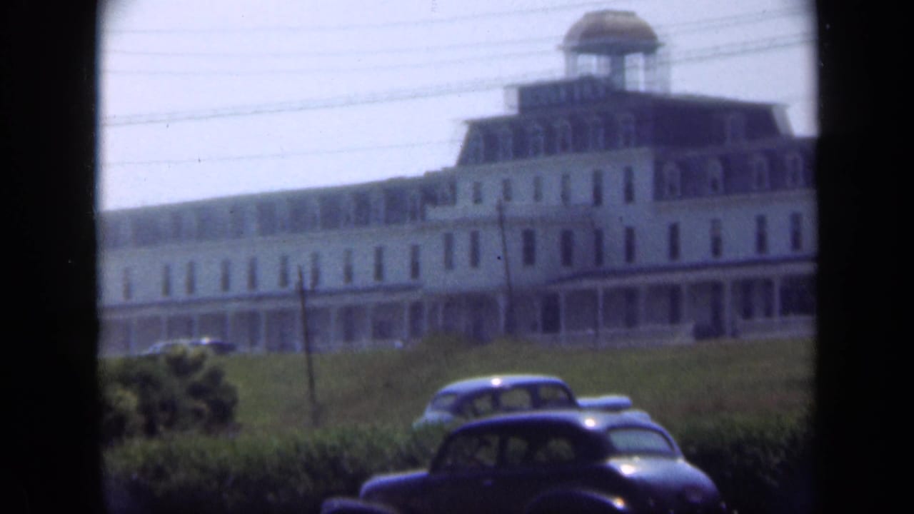 Large white hotel on a grassy rise with vintage cars in the foreground.