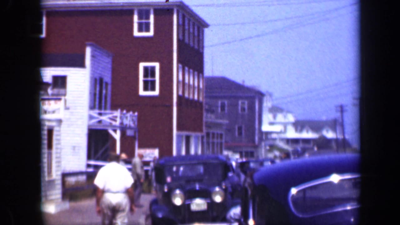 Pedestrians and vintage cars on a street beside multi-story buildings.