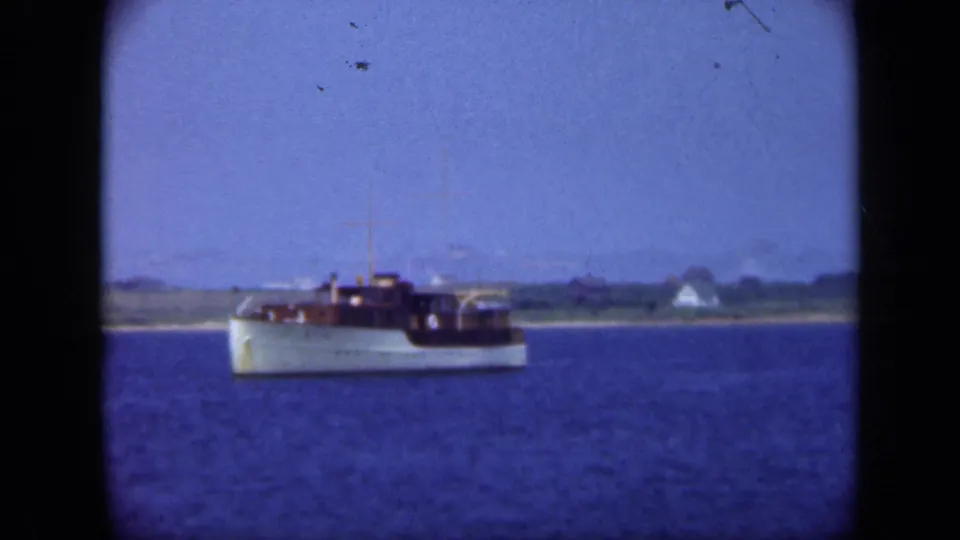 Classic white-and-brown motor yacht crossing calm water off Long Island, with low land and small buildings in the background.