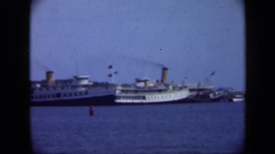 A Long Island harbor frame shows large steamships moored across blue water, with smoke visible above a funnel.