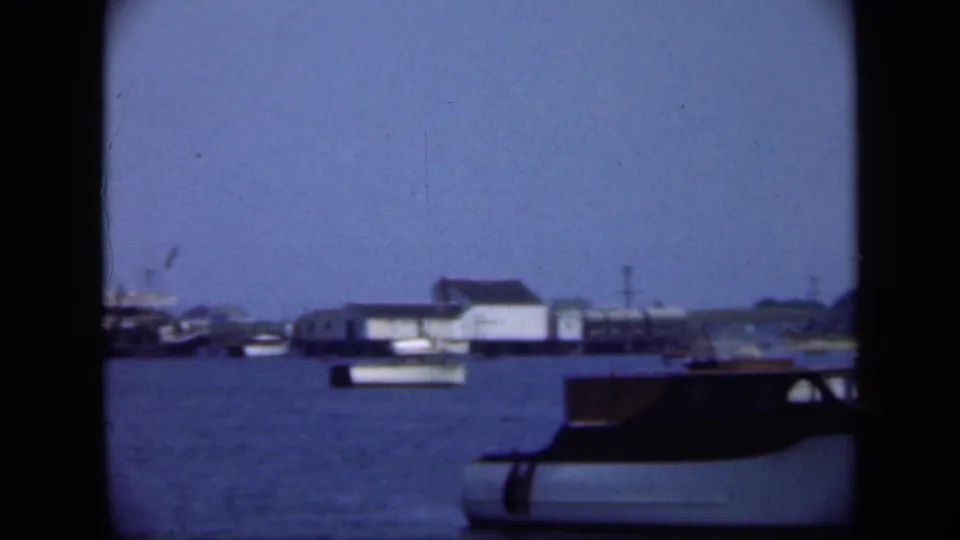 Distant Long Island harbor scene with low waterfront buildings, small boats, and the edge of a dark foreground object.