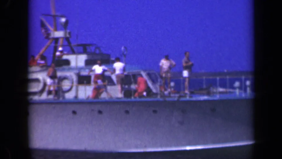 Mid-century home-movie frame of a small passenger boat deck with people, railings, lifebuoys, and a raised structure under a blue sky.