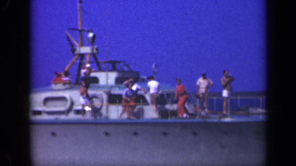 1948 Long Island boating frame showing passengers gathered on deck near railings, lifebuoys, and a mast-like structure in bright blue daylight.