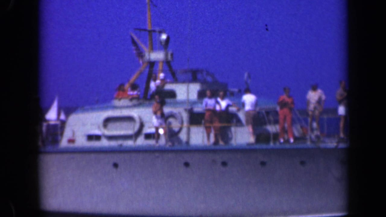 White boat deck with several people, railings, mast structure, flag, and blue sky.