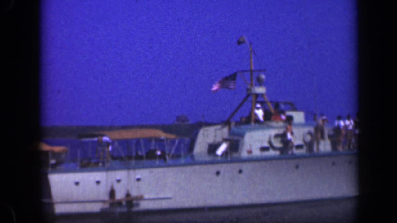 Side view of the Long Island pleasure boat, with people on deck, a small cabin structure, canopy, flag, and shoreline beyond.
