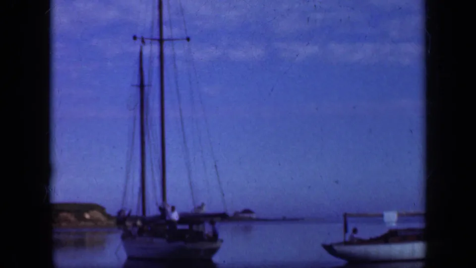 1948 Long Island waterfront frame showing a sailboat with tall masts on calm water, with shoreline and blue sky in the distance.