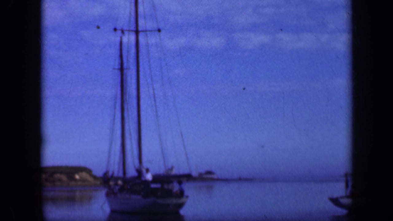 Moored sailboat with lowered sail on blue water near a low shoreline.