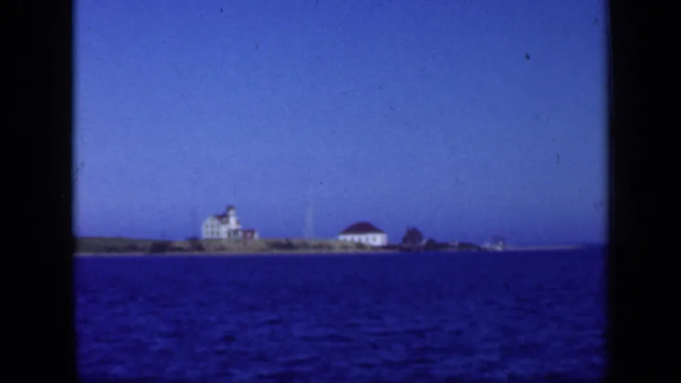 1948 Long Island shoreline view with several distant waterfront buildings, red roofs, blue bay water, and soft 8mm texture.