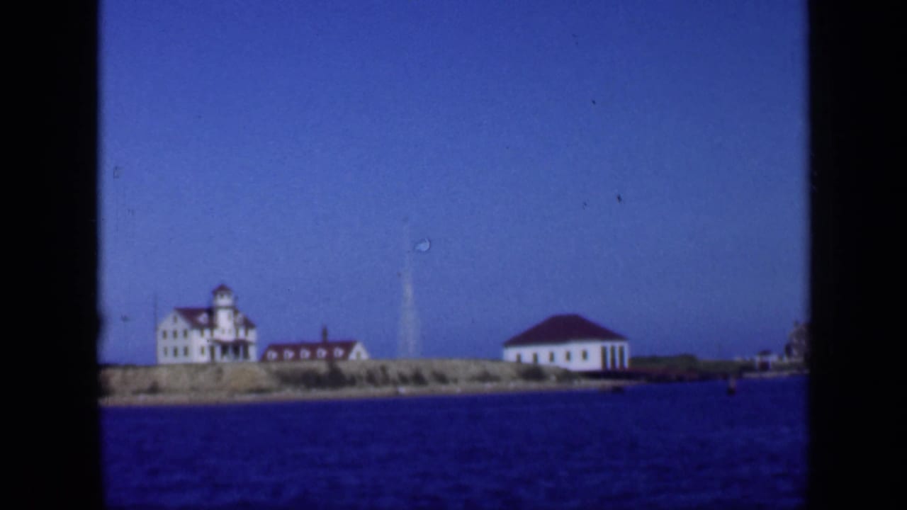 Long Island shoreline filmed from the water, with white buildings, red roofs, and a lighthouse-like tower visible.