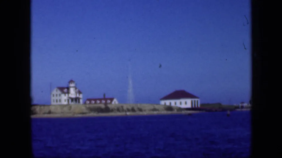 8mm frame of a Long Island coastal shoreline with white red-roofed buildings and a tower-like structure across the water.