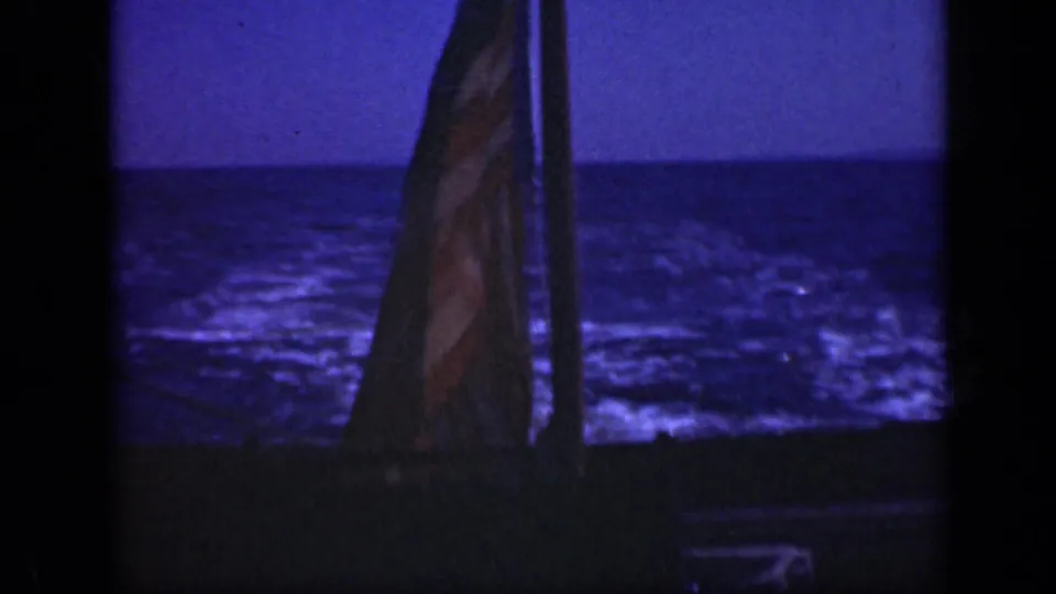 View from the stern of a small boat, with a flag silhouetted against open water in low blue light.