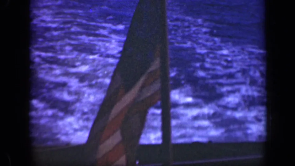 The U.S. flag fills the right side of a 1948 Long Island-area shipboard frame, with textured ocean water behind it.