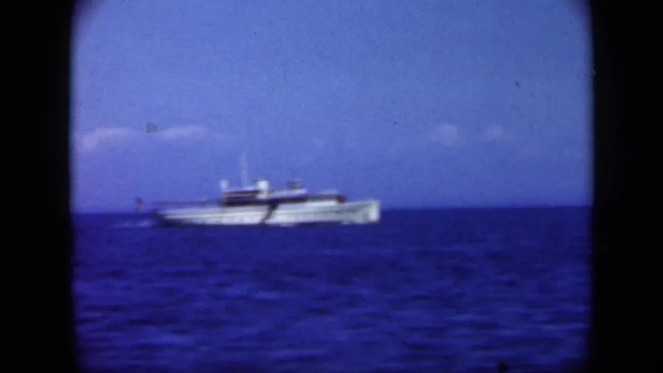 White boat moving across blue Long Island water in 1948 8mm footage, with low horizon and cloudy sky behind it.