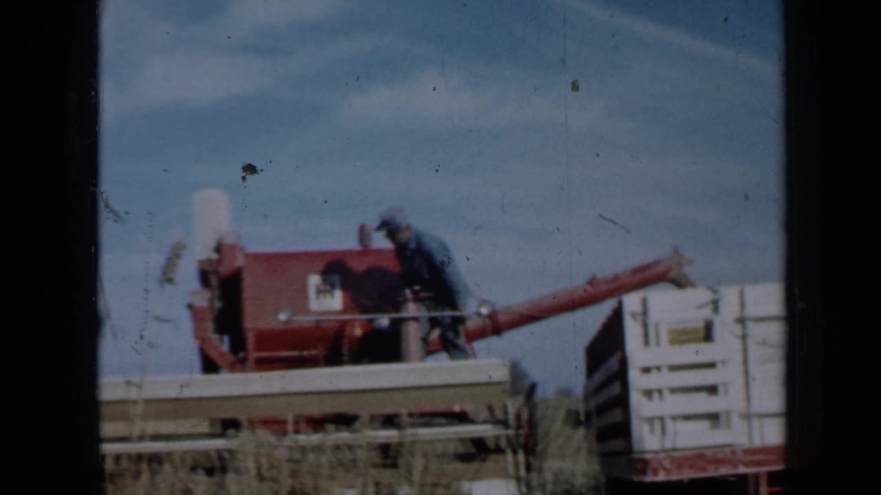 Farmer seated on a red combine beside a white wooden grain box in a field.