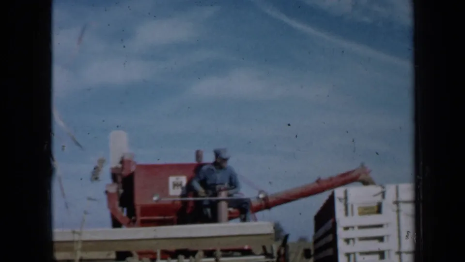 The 8mm frame shows the farmer on the combine from a slightly shifted angle, with dry stalks in the foreground and grain box nearby.