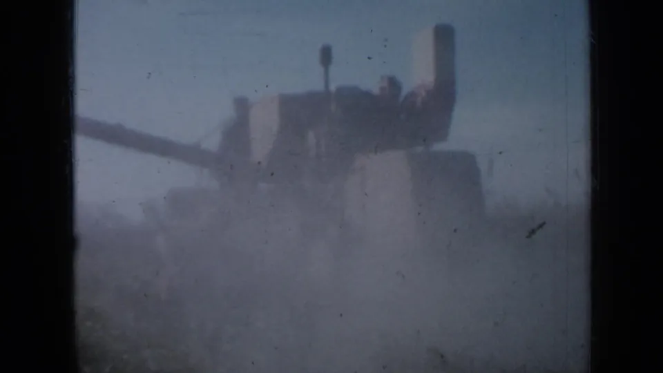 A grain harvester appears through a dense dust cloud in the Minnesota field, with its long auger extending left.