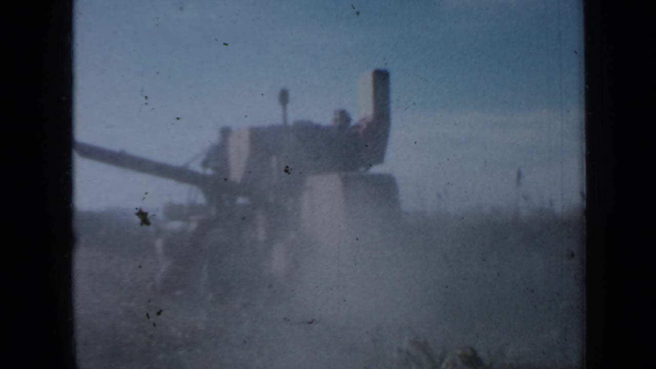 1958 Minnesota 8mm frame showing a distant tractor or harvester working through tall crops under a hazy sky.