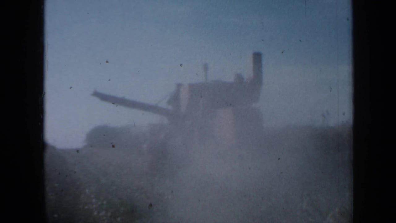 1958 Minnesota home-movie frame with a distant farm machine moving near tall crops in a hazy field.