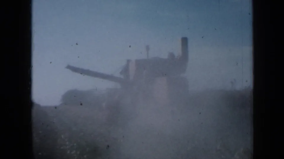 1958 Minnesota home-movie frame with a distant farm machine moving near tall crops in a hazy field.