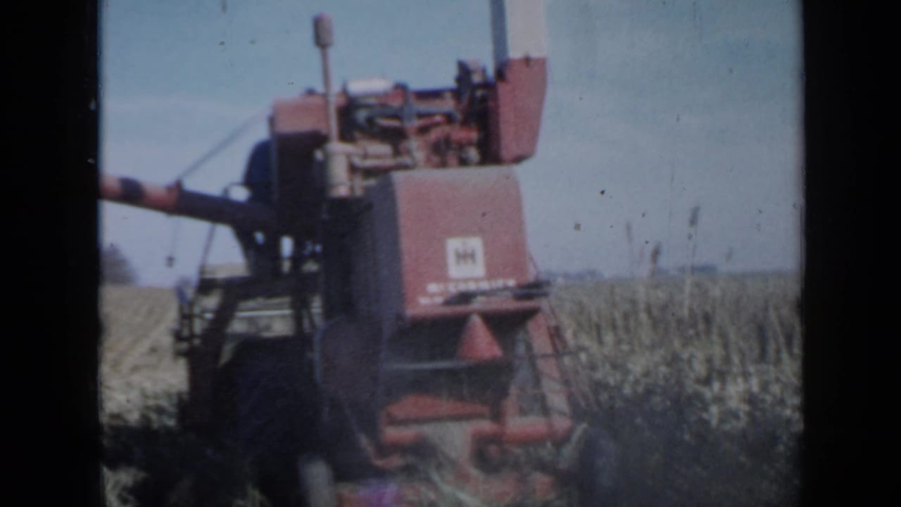 1958 Minnesota 8mm frame showing a red combine harvester moving through corn with open field rows and blue sky behind it.
