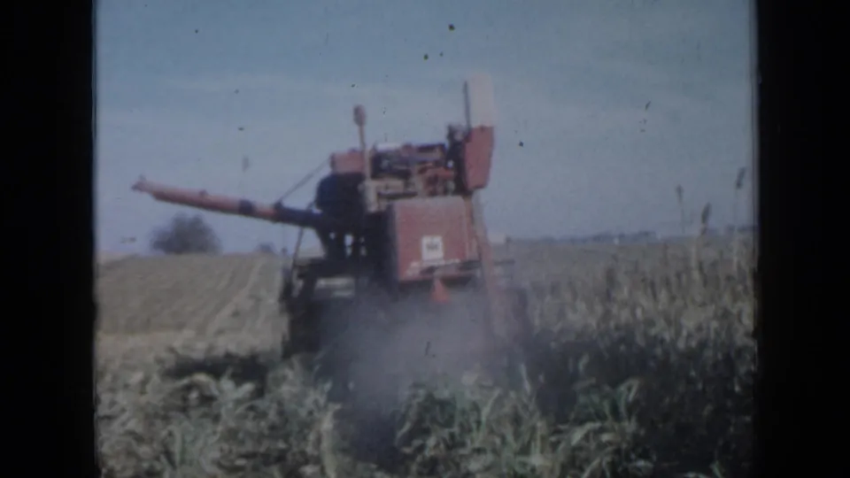 1958 Minnesota 8mm frame of a red combine harvesting corn, with dust softening the rear of the machine and field rows beyond.