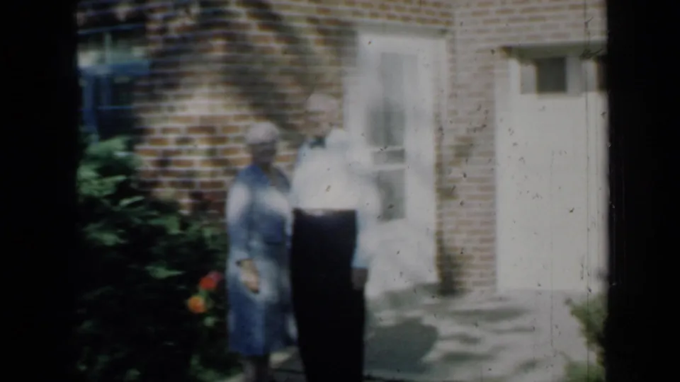 1958 home-movie frame showing an older couple standing outside a brick house entrance in Minnesota.